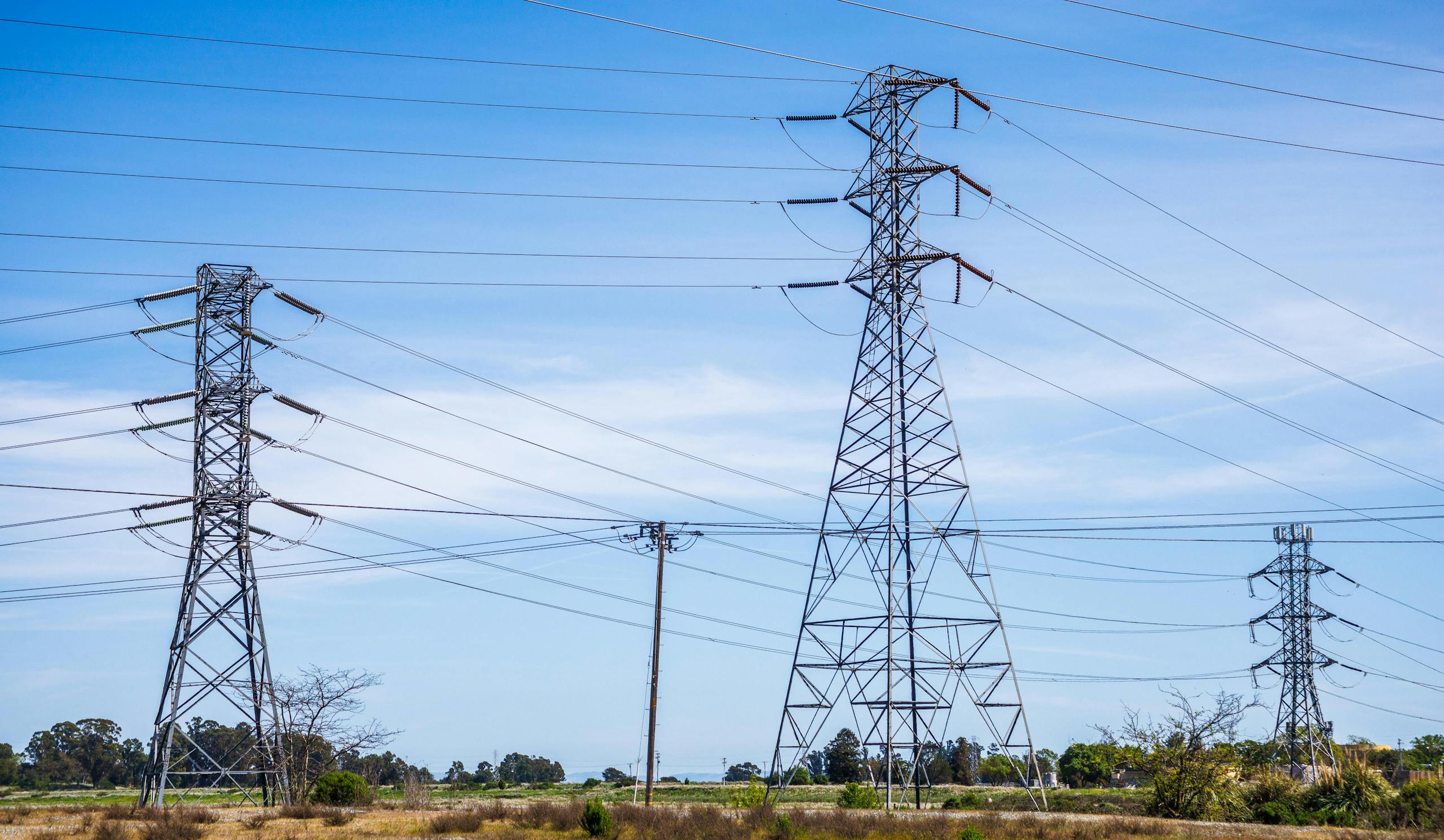 High voltage electric pylons in a field under a clear blue sky demonstrate power transmission infrastructure.